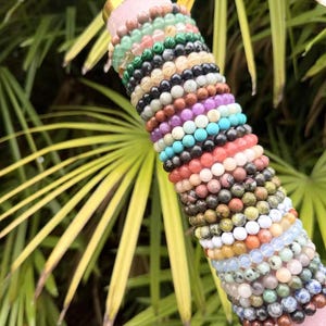 Könnte beinhalten: Eine Sammlung von Perlenarmbändern in verschiedenen Farben, darunter Grün, Schwarz, Braun, Lila und Türkis. Die Armbänder sind gestapelt und zeigen eine Vielzahl von Natursteinperlen. Der Hintergrund zeigt grünes Laub.