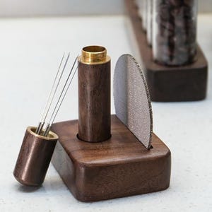 Puede incluir: Un juego de herramientas de distribución de café espresso de madera. El juego incluye una base de madera marrón oscuro, una herramienta de distribución con agujas, una pantalla de portafiltro y un soporte de madera con una parte superior de latón. Las herramientas están diseñadas para la preparación de café.