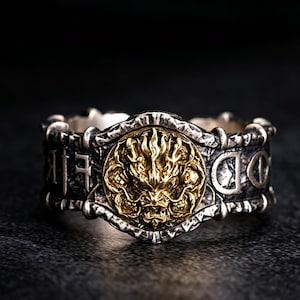 May include: A silver-toned ring with a gold-colored dragon design in the center. The ring has a detailed, textured band with engraved symbols. The dragon is the focal point, with intricate details and a bold presence.