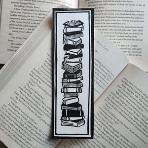 Puede incluir: Marcapáginas en blanco y negro con una pila de libros. La ilustración muestra una alta pila de libros de varios tamaños y grosores, ideal para lectores. El marcapáginas es rectangular con un borde negro.