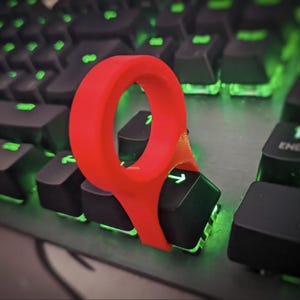 May include: A red keyboard key puller tool is placed over a black keyboard key with a white arrow. The keyboard has black keys with green backlighting. The tool is made of a flexible material.