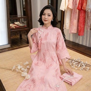 May include: A light pink, traditional Chinese dress with a mandarin collar and floral embroidery. The dress features three-quarter sleeves and decorative frog closures. A matching pink fabric is folded on the floor.