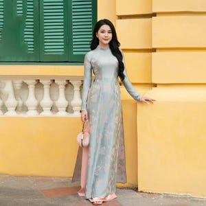 以下が含まれることがあります： 花柄のロング丈ライトブルーのドレス。長袖で、ハイネック。スリットからピンクのインナースカートが見える。手に小さなピンクの丸いハンドバッグを持っている。