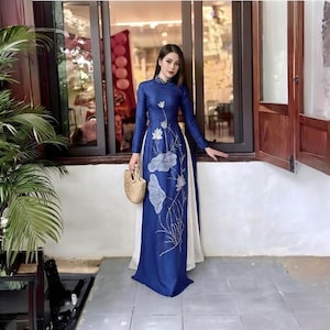 May include: A long, royal blue dress with white accents and a floral design. The dress features long sleeves and a high collar. A woven straw handbag is held in the hand.