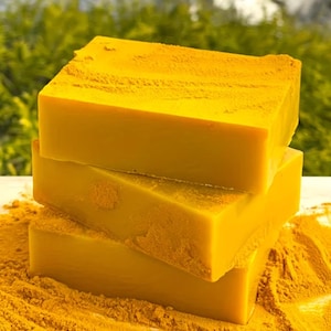 May include: Three bright yellow soap bars stacked on top of each other, with a textured surface, and a bed of yellow powder. The soaps are square and have a smooth, solid appearance, suggesting natural ingredients.