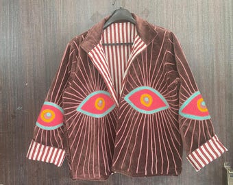 May include: Brown velvet jacket with embroidered eye designs. The jacket features a striped lining and cuffs, with pink and blue accents. The eye designs are on the front and sleeves.