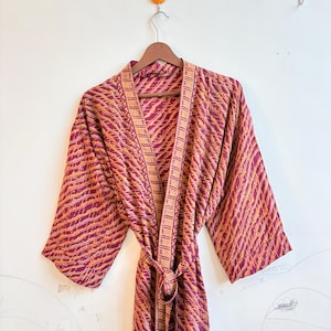 May include: A burgundy robe with an orange, striped pattern, hanging on a wooden hanger. The robe has a V-neck and a matching belt. The fabric appears lightweight and flowing, suitable for lounging or as a cover-up.