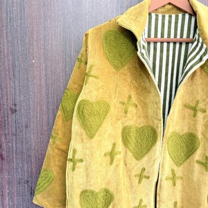 Peut inclure: Veste jaune moutarde avec des cœurs et des croix verts brodés. La veste a un col et une doublure rayée verte et blanche. La veste est suspendue à un cintre en bois.