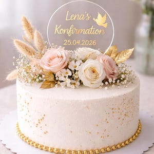 Könnte beinhalten: Eine weiß glasierte Torte, verziert mit rosa und weißen Rosen, kleinen weißen Blüten und goldenen Akzenten. Ein klarer Aufsatz zeigt "Lena's Konfirmation 25.04.2026" mit einer goldenen Taube. Die Torte hat einen goldenen Perlenrand.