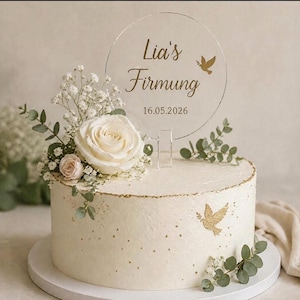 May include: A round, white frosted cake decorated with white roses, greenery, and gold accents. A clear acrylic topper reads "Lia's Firmung" and the date 16.05.2026. A dove design is also featured. The cake is on a white cake stand.
