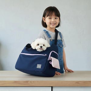 May include: A navy blue pet carrier with a light pink zip and trim, holding a small white dog. The carrier has a shoulder strap and a small pocket. The dog is looking out of the carrier. The carrier is on a wooden surface.