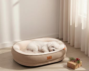 Puede incluir: Una cama para perros ovalada de pana beige con interior crema. Un perro blanco pequeño duerme plácidamente dentro. Una pequeña caja de regalo con vegetación se coloca al lado. Un accesorio acogedor para mascotas.