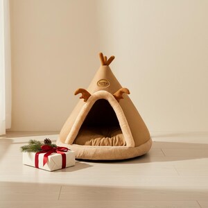 May include: A tan pet bed shaped like a teepee with brown antler accents and a small logo. The bed has a soft, plush interior and a rounded base. A wrapped gift with a red bow and greenery sits nearby.