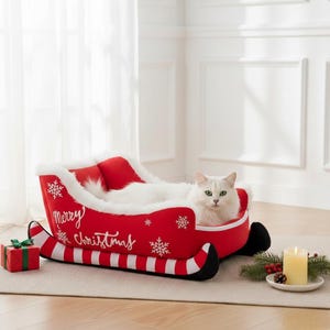 May include: A white cat rests in a red Christmas sleigh-shaped pet bed. The sleigh features white trim, snowflakes, and the words "Merry Christmas." A small gift and candle are present. The bed is on a neutral rug.