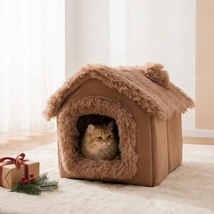 以下が含まれることがあります： 小さな家の形をした茶色の猫用ハウス。入り口と屋根の周りにふわふわのフェイクファーのトリムが付いています。ふわふわの猫が中にいます。白いラグの上に家があります。