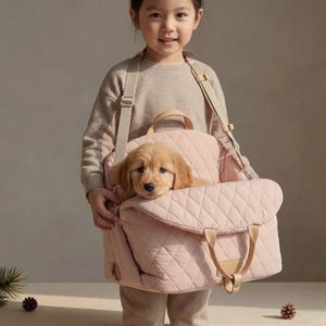 Op de afbeelding: Een lichtroze gewatteerde draagtas voor huisdieren met een golden retriever puppy erin. De tas heeft beige riemen en handvatten. De puppy kijkt naar de camera. De tas wordt door een persoon vastgehouden.