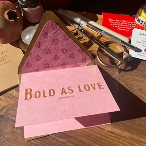 Op de afbeelding: Een verzameling briefpapierartikelen, waaronder een roze kaart met de tekst "Bold as Love" en een citaat van Jimi Hendrix. Andere items zijn een visitekaartje, een schaar en decoratieve tape. De items zijn gerangschikt op een houten oppervlak.