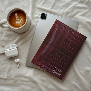 May include: A burgundy leather tablet case with a crocodile skin pattern, personalised with the name "Natalie Richards" in white script. The case rests on a silver tablet, next to a latte and white wireless earbuds.