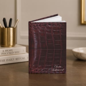 May include: A vertical notebook with a dark burgundy, textured crocodile-patterned cover. The front cover features the name Claire Underwood embossed in a delicate, light-colored script font. The notebook is slightly open, revealing white pages.