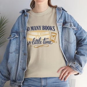 May include: Beige t-shirt with the text "So Many Books So Little Time" in navy blue and gold. The design includes stacked books and an hourglass. The person is wearing a light blue denim jacket.