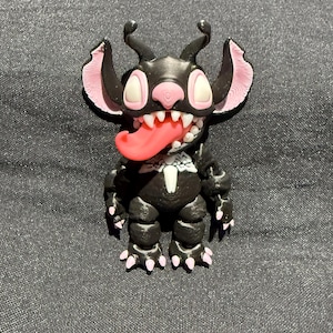 Puede incluir: Una figura negra de un personaje de dibujos animados con orejas rosas, una nariz rosa y una lengua roja. El personaje tiene dientes blancos y un dise&ntilde;o blanco en el pecho. La figura est&aacute; sobre una superficie gris.