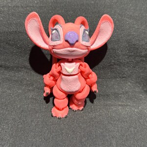 Puede incluir: Figura impresa en 3D de un personaje de dibujos animados rosa con orejas grandes, una nariz p&uacute;rpura y rasgos faciales detallados. La figura tiene un peque&ntilde;o emblema blanco en el pecho y est&aacute; de pie. La figura mide aproximadamente 10 cm de alto.