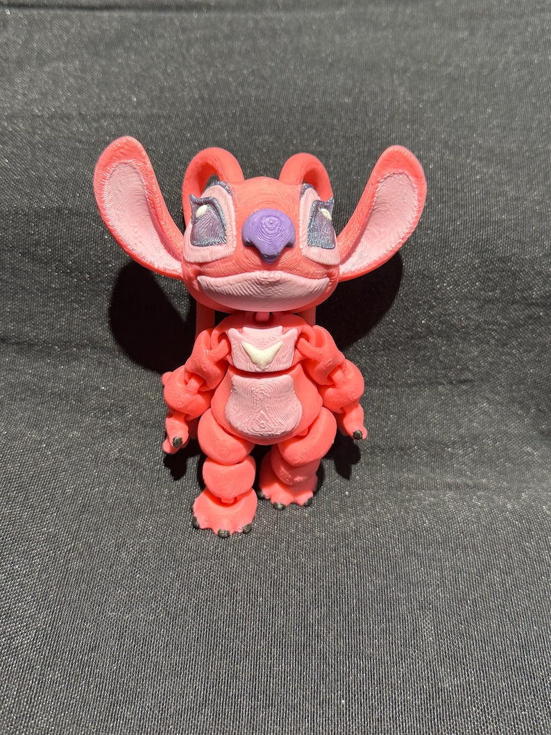 Puede incluir: Figura impresa en 3D de un personaje de dibujos animados rosa con orejas grandes, una nariz p&uacute;rpura y rasgos faciales detallados. La figura tiene un peque&ntilde;o emblema blanco en el pecho y est&aacute; de pie. La figura mide aproximadamente 10 cm de alto.
