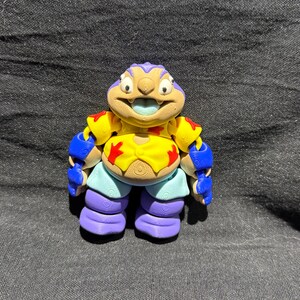 Puede incluir: Una figura colorida y estilizada de un personaje de dibujos animados. La figura tiene una cara bronceada con una lengua azul, una cabeza morada y una armadura azul y amarilla. La figura lleva pantalones cortos azul claro y botas moradas.