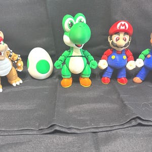 Puede incluir: Un conjunto de cuatro figuras de plástico que incluye un Bowser marrón y beige, un huevo blanco con manchas verdes, un Yoshi verde, una figura de Mario con gorra roja y peto azul, y una figura de Luigi con gorra verde y peto azul.