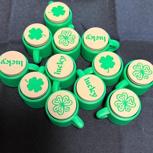 Può includere: Una collezione di oggetti verdi a forma di tazza con coperchi marrone chiaro. Ogni coperchio presenta un trifoglio verde o la parola "lucky" in verde. Le tazze sono disposte su una superficie scura, evocando il giorno di San Patrizio.