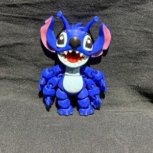 Puede incluir: Una figura articulada azul de un personaje alien&iacute;gena de dibujos animados con orejas grandes, una amplia sonrisa y m&uacute;ltiples brazos. La figura tiene detalles en rosa en las orejas y la boca, y est&aacute; posada sobre un fondo oscuro.