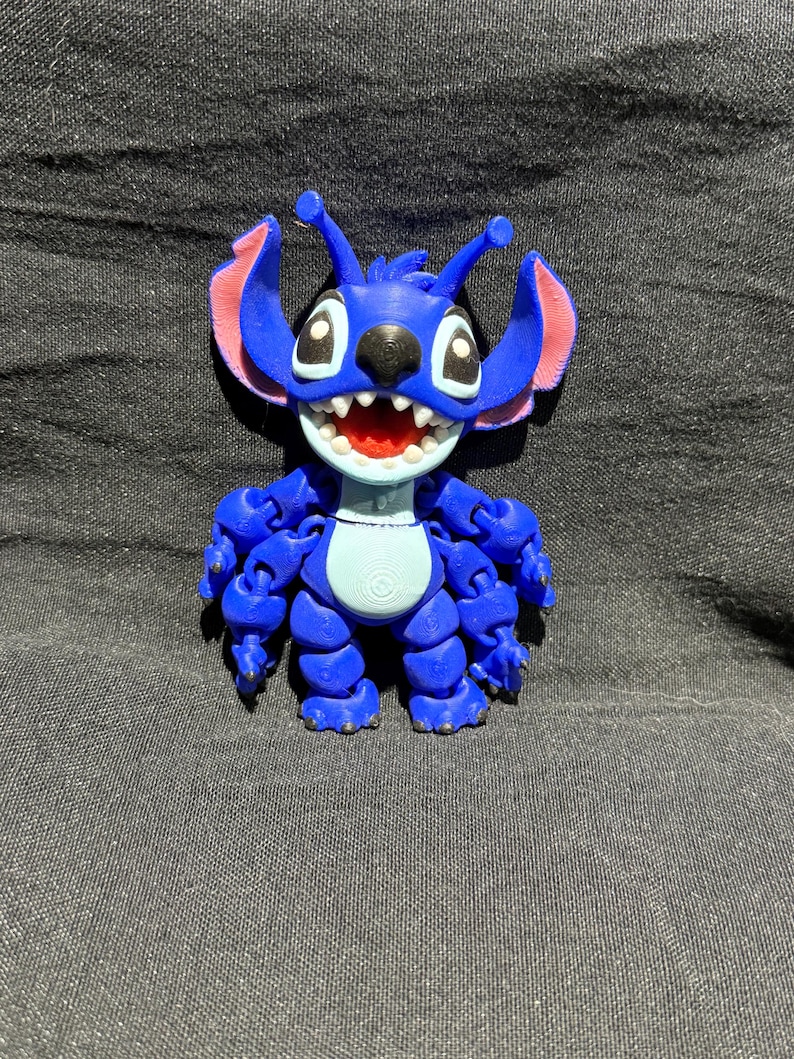Puede incluir: Una figura articulada azul de un personaje alien&iacute;gena de dibujos animados con orejas grandes, una amplia sonrisa y m&uacute;ltiples brazos. La figura tiene detalles en rosa en las orejas y la boca, y est&aacute; posada sobre un fondo oscuro.