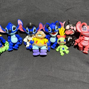 Puede incluir: Una colecci&oacute;n de figuras de personajes coloridos, que incluyen criaturas azules, verdes y rojas con orejas grandes y caras expresivas. Las figuras est&aacute;n dispuestas sobre una superficie gris oscuro.