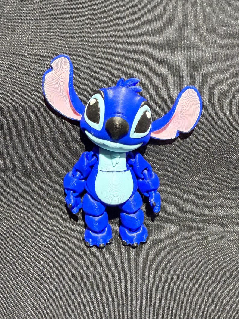 Puede incluir: Una figura articulada azul del personaje Stitch de la pel&iacute;cula de Disney Lilo & Stitch. La figura tiene orejas grandes con interiores rosas, ojos negros y una barriga azul claro. La figura est&aacute; sobre una superficie gris oscuro.