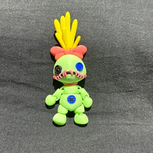 Puede incluir: Una figura articulada impresa en 3D de un personaje de dibujos animados. La figura es principalmente verde claro con un lazo rosa y detalles amarillos en forma de cabello. Tiene detalles cosidos en negro y ojos azules. La figura est&aacute; sobre una superficie gris oscuro.