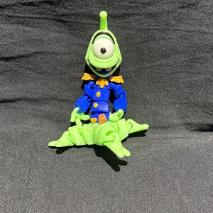 Puede incluir: Un juguete de alien&iacute;gena verde con un solo ojo, que lleva un uniforme azul con detalles dorados. El alien&iacute;gena est&aacute; posado sobre una base verde claro y estilizada. El juguete est&aacute; sobre un fondo gris oscuro.