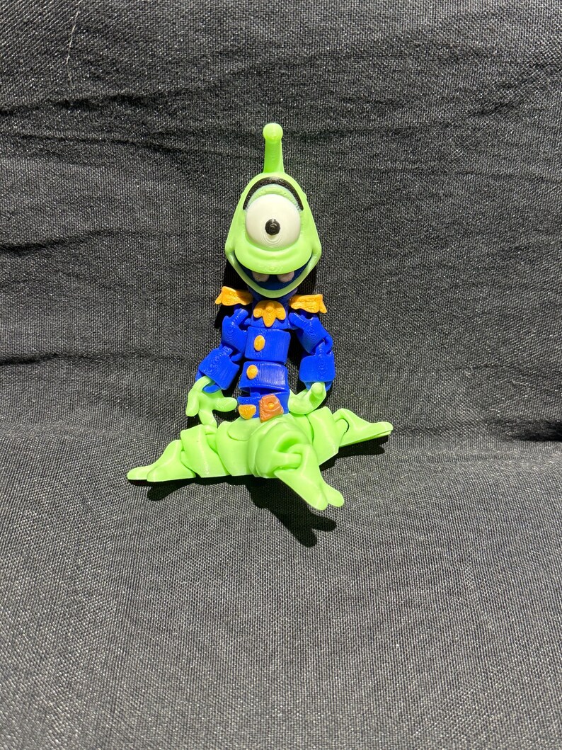 Puede incluir: Un juguete de alien&iacute;gena verde con un solo ojo, que lleva un uniforme azul con detalles dorados. El alien&iacute;gena est&aacute; posado sobre una base verde claro y estilizada. El juguete est&aacute; sobre un fondo gris oscuro.