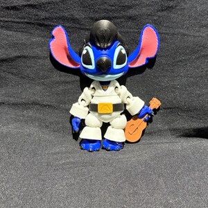 Puede incluir: Una figura azul y blanca del personaje Stitch, vestida con un mono blanco con cintur&oacute;n dorado y peinado negro. La figura sostiene una peque&ntilde;a guitarra marr&oacute;n. Las orejas son rosas.