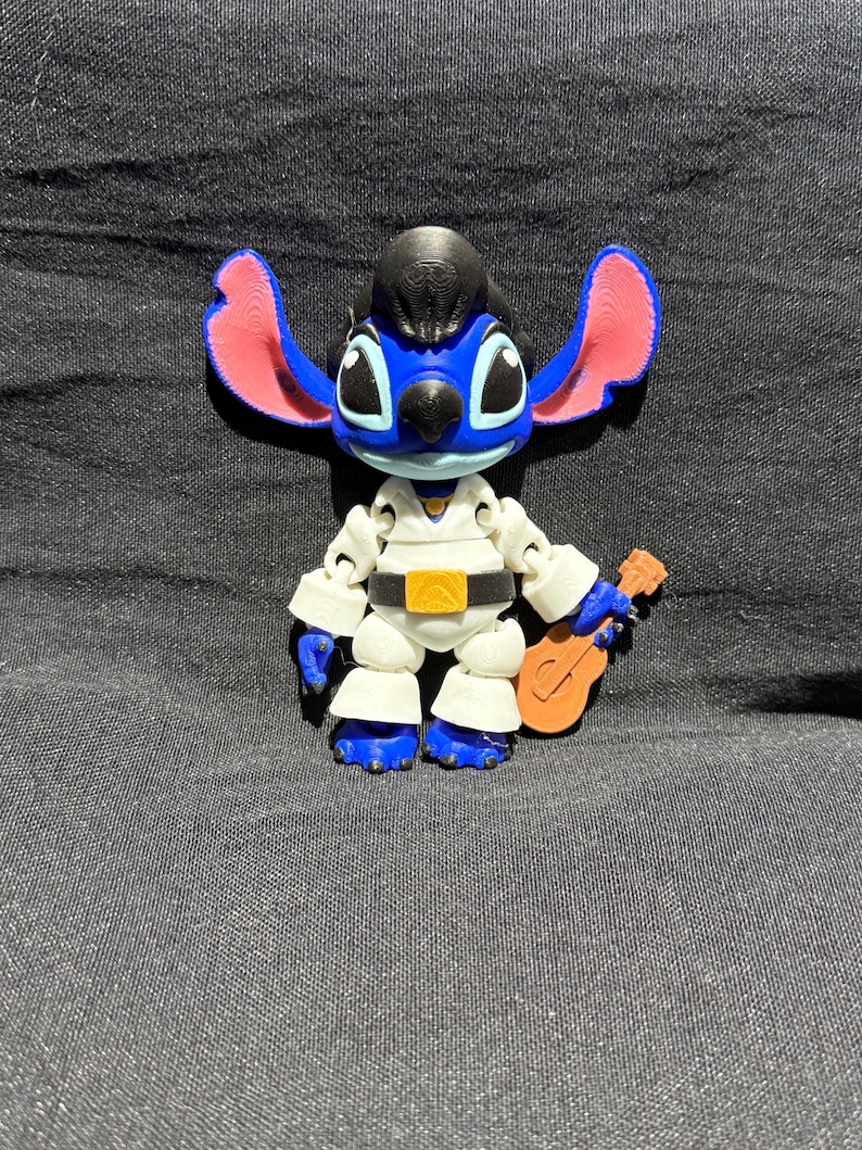 Puede incluir: Una figura azul y blanca del personaje Stitch, vestida con un mono blanco con cintur&oacute;n dorado y peinado negro. La figura sostiene una peque&ntilde;a guitarra marr&oacute;n. Las orejas son rosas.