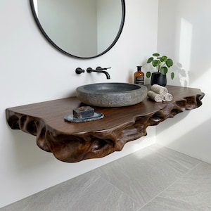 Puede incluir: Un mueble de baño flotante de madera oscura con un borde natural ondulado. Incluye un lavabo de piedra gris, un grifo negro mate montado en la pared, una pequeña jabonera con una pastilla de jabón oscura y tres toallas de color beige enrolladas.