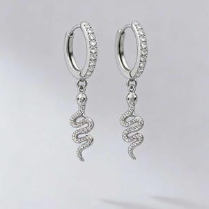 Może przedstawiać: Para srebrnych kolczyków koła z wisiorkiem w kształcie węża. Koła są inkrustowane małymi, przezroczystymi kamieniami. Każdy kolczyk ma szczegółowy urok węża, również ozdobiony przezroczystymi kamieniami, tworząc efekt połysku.