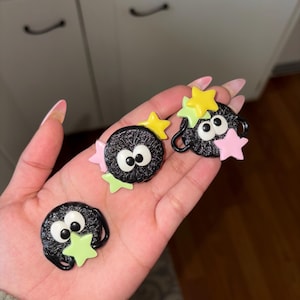 Puede incluir: Tres adornos negros y redondos, tipo caricatura, con ojos blancos y detalles de estrellas en amarillo, rosa y verde. Los adornos se sostienen en una mano.