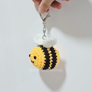 May include: A crocheted bee keyring with yellow and black stripes, white wings, and a silver-coloured clasp. The bee has small black eyes and is suspended against a white background. The text "@crochetmusings" is visible.