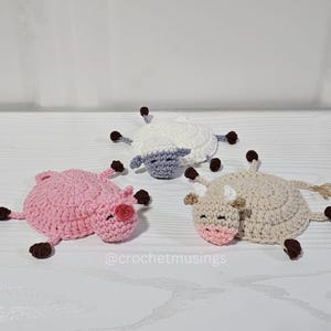 May include: Three handmade crochet farm animal toys: a pink pig, a white sheep, and a tan cow. Each animal has brown accents and a smiling face. The toys are lying on a white surface.