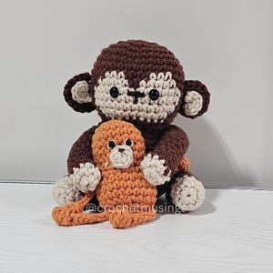 Op de afbeelding: Een handgemaakte gehaakte apenpop in bruin, beige en oranje. De grotere aap houdt een kleinere aap vast. De poppen hebben zwarte veiligheidsoogjes en staan op een wit oppervlak. De tekst "@crochetmusing" is zichtbaar.