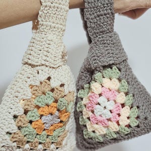 May include: Two handmade crochet bags are held up. One is off-white with a floral pattern in orange, green, and brown. The other is gray with a pink, white, and green floral pattern. The bags have a square base and a handle.