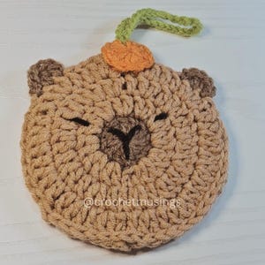 Könnte beinhalten: Ein gehäkeltes Capybara-Gesicht in Brauntönen, mit kleinen Ohren und geschlossenen Augen. Es hat einen kleinen orangefarbenen Kürbis und eine grüne Schlaufe oben. Der Text "@crochetmusings" befindet sich unten.
