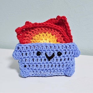 May include: A handmade crochet dumpster toy in light blue with a smiling face and a red, orange, and yellow flame. The toy is sitting on a white surface. The text "@crochetinshps" is visible on the front.