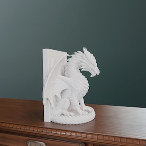 Op de afbeelding: Een witte drakenboekensteun. De draak zit met uitgespreide vleugels naast een verticale steun. De sculptuur heeft gedetailleerde schubben en een felle uitdrukking. De boekensteun staat op een houten oppervlak.