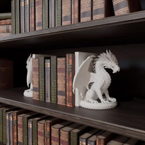 Op de afbeelding: Witte drakenboekensteunen met gedetailleerde vleugels en schubben, geplaatst op een donkere houten plank. De drakenboekensteunen staan tussen een verzameling antieke boeken met verschillende kleuren en texturen. De plank maakt deel uit van een boekenkast.